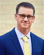 Headshot of Supt. Micah Hill wearing a blue suit, yellow tie, and glasses.